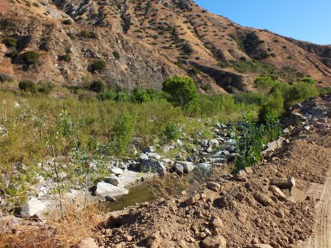 2003 Ford Crown Victoria - shallow rocky stream cuts through a dry canyon flanked by graded dirt and green growth.