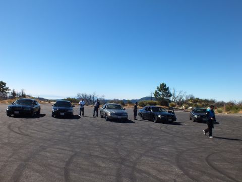 2003 Ford Crown Victoria - Drivers stand among parked full-size sedans on skid-marked pavement on a hilltop.