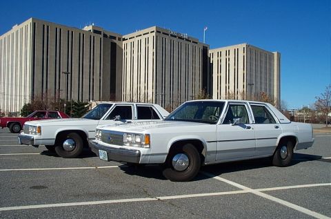 1991 Ford LTD Crown Victoria - Two white Ford Crown Victorias and a red sedan are parked in a lot with a large building in the background.
