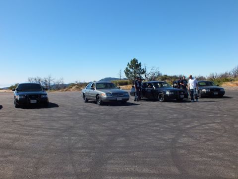 2003 Ford Crown Victoria - People cluster beside dark sedans on a skid-marked pad above the hills.