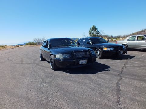 2003 Ford Crown Victoria - Pair of murdered-out Panther-platform sedans on hilltop asphalt, push bar on lead car, hazy valley behind