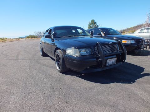 2003 Ford Crown Victoria - Front-quarter of blacked-out Panther sedan with grille guard and California plate, second car at right