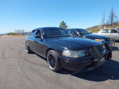 2003 Ford Crown Victoria - Lowered blacked-out sedan at low angle, brush guard up front, dead-tree hillside behind