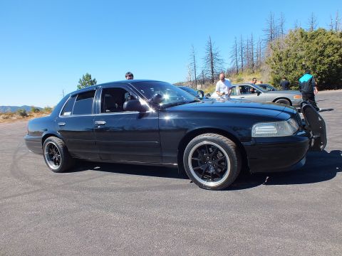 2003 Ford Crown Victoria - Lowered blacked-out sedan with grille guard, group of people and silver car in background