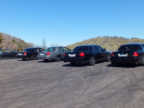 2003 Ford Crown Victoria - Black and silver Crown Victorias lined up rear-quarter, dry hillside and clear sky behind