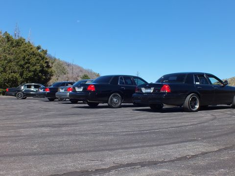 2003 Ford Crown Victoria - Murdered-out Panther sedans lined up low to ground seen from rear, hillside in background