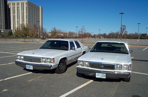 1991 Ford LTD Crown Victoria - Two white 1991 Ford Crown Victorias are parked side-by-side in an empty lot under a clear blue sky.