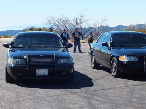 2003 Ford Crown Victoria - Two black sedans face camera as people watch; one wears a police brush guard