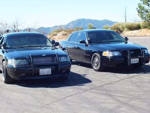 2003 Ford Crown Victoria - Pair of blacked-out Ford and Mercury sedans on asphalt with green mountains behind