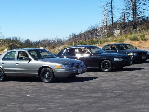 2003 Ford Crown Victoria - Trio of full-size sedans angled on cracked asphalt beneath a scorched ridge