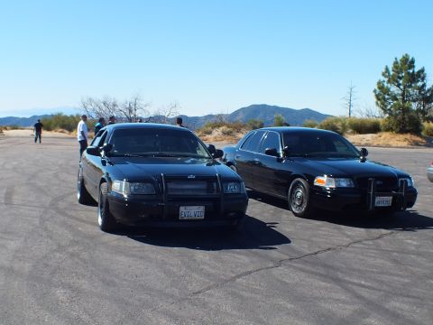 2003 Ford Crown Victoria - Two blacked-out sedans on a lot with onlookers and mountains in the background