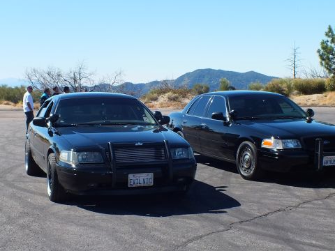 2003 Ford Crown Victoria - Two dark full-size Ford and Mercury sedans parked angled before brushy hills