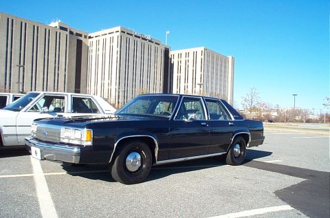 1991 Ford LTD Crown Victoria - A dark blue sedan is parked in a lot with a white car beside it, under a clear blue sky.