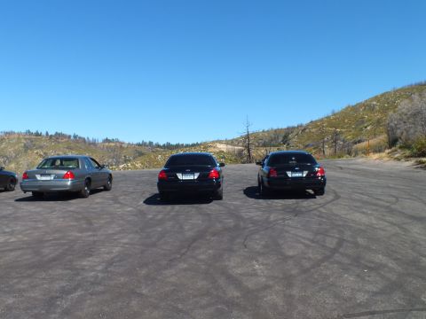 2003 Ford Crown Victoria - Rear view of black and silver Ford sedans parked on a hillside lot overlooking burned terrain