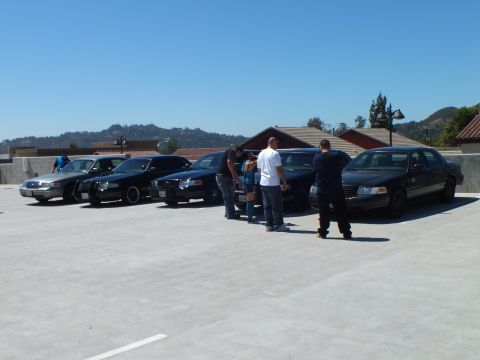 2003 Ford Crown Victoria - Group inspects a row of dark full-size sedans on a rooftop deck below dry hills