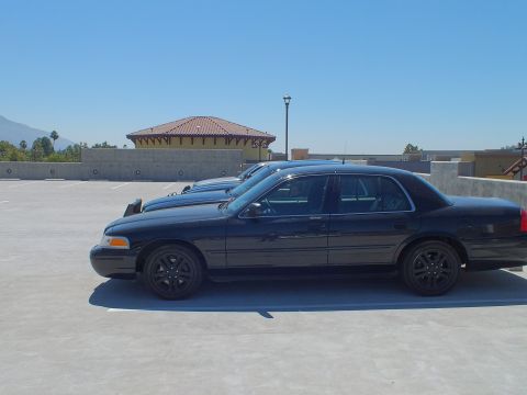 2003 Ford Crown Victoria - Row of blacked-out Crown Victoria sedans on an open-air parking structure, mountains behind.