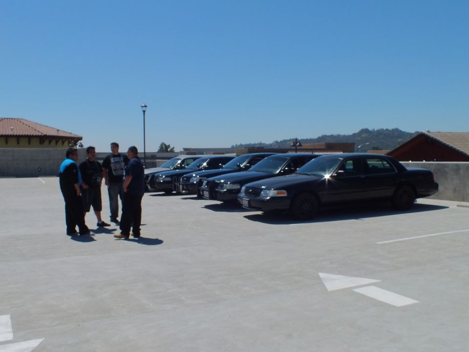 Group of people beside black Ford Crown Victoria sedans on rooftop parking deck