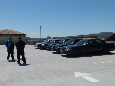 2003 Ford Crown Victoria - Black Crown Victoria sedans angled in a row with two bystanders on a parking structure.