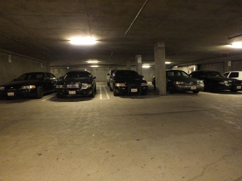 2003 Ford Crown Victoria - Lineup of dark sedans beside a support column on a basement parking level.