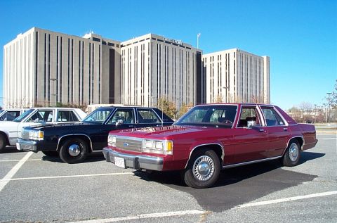 1991 Ford LTD Crown Victoria - Three sedans are parked in a lot with a large building in the background.