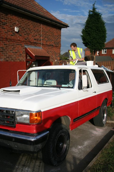 1989 Ford Bronco - A person is on the roof of a red and white 1989 Ford Bronco parked outside a brick house.
