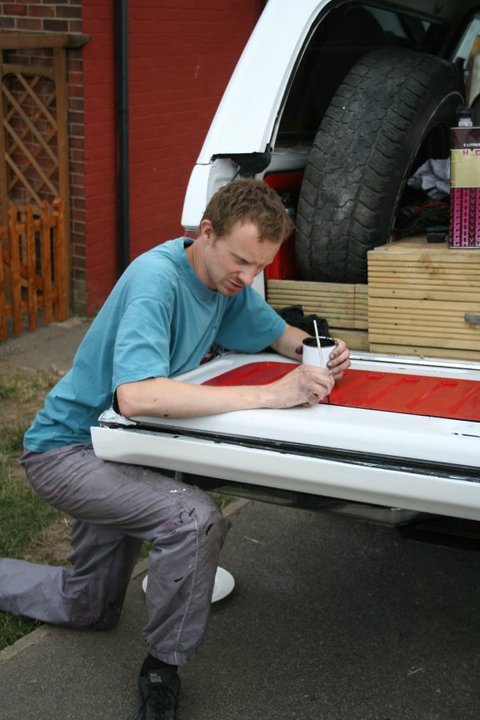 1989 Ford Bronco - A man kneels to paint a red stripe onto the white tailgate of a vehicle, with a spare tire visible in the cargo area.