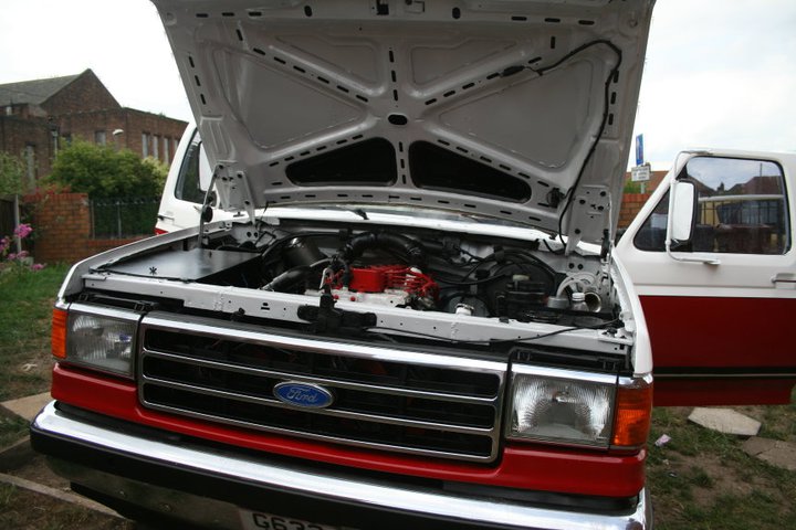 Ford F-Series truck with hood open showing engine. Red stripe on lower body. Another white vehicle behind it