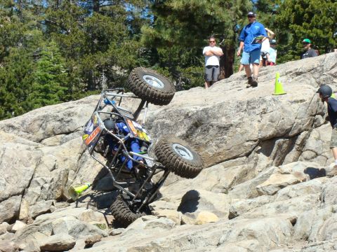 1989 Ford Bronco - An off-road buggy struggles on a steep rocky terrain during a competition, with onlookers observing the challenging obstacle.