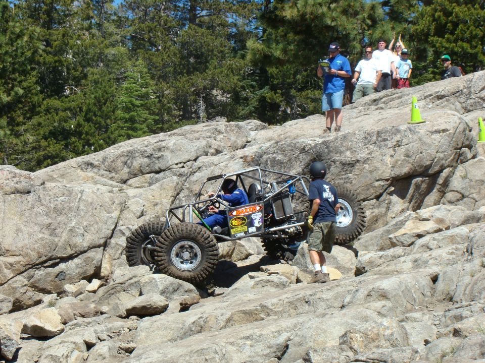 Off-road buggy with roll cage climbing rocky terrain, driver in blue suit