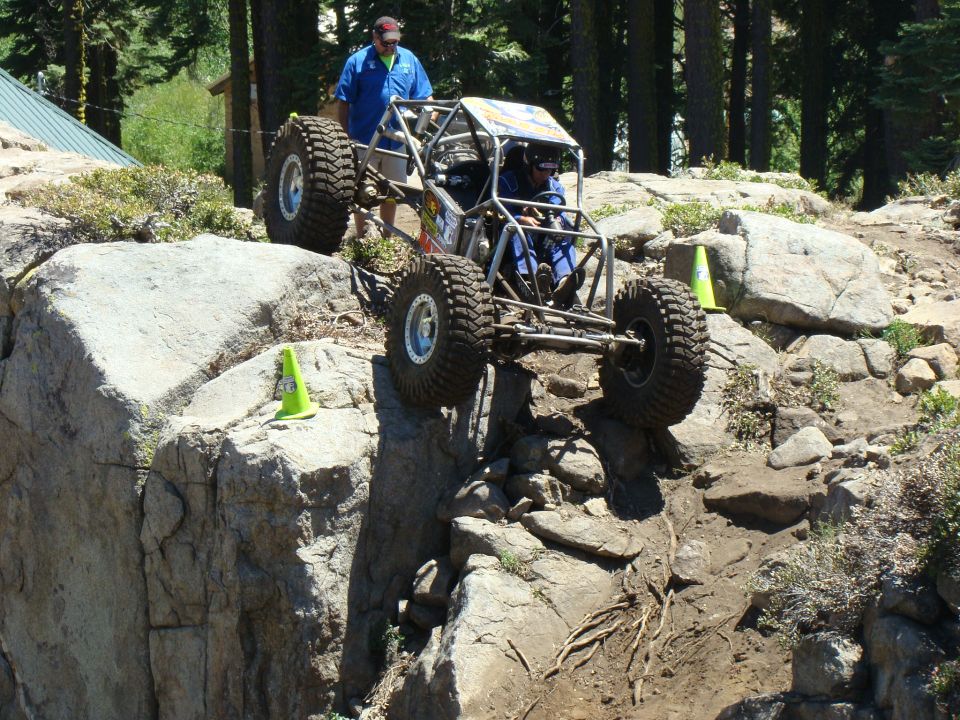 Custom off-road buggy with large tires navigating a steep rocky incline, driver visible