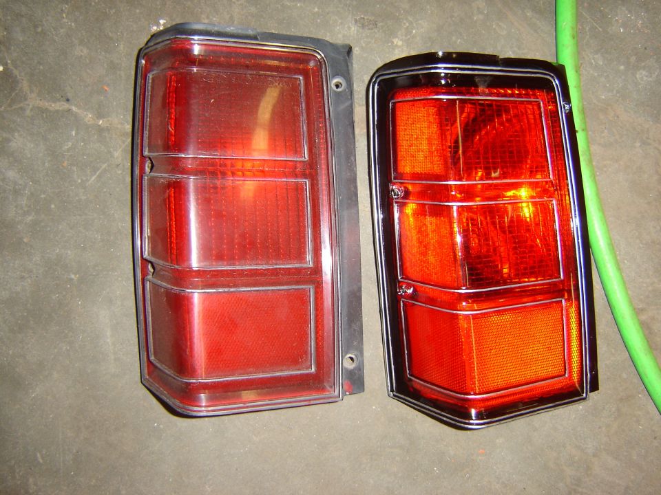 Two red tail lights, one with a chrome bezel and one with a black bezel, against a concrete floor