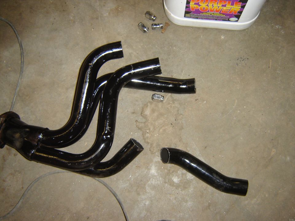 Black exhaust manifold with four pipes, cut off at the ends, lying on a concrete floor next to a bottle of Purple Power cleaner