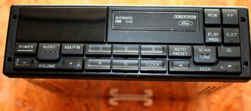 Car stereo with Ford logo, cassette deck, and numerous buttons including POWER, AUDIO, AM/FM, VOLUME, CLOCK, SCAN, TUNE, REW, FF, PLAY, PROG, and EJECT