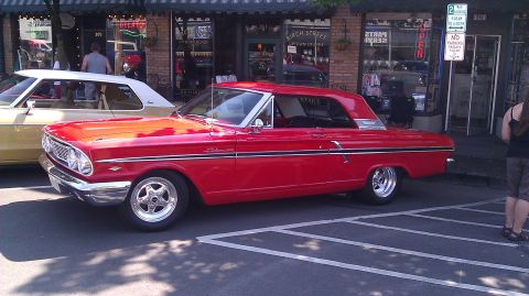 1964 Ford Fairlane - A red 1964 Ford Fairlane coupe is parked next to another vehicle in front of a storefront.