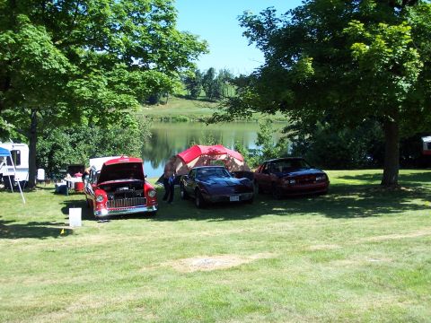 1975 Ford Ranchero - Three cars are parked on the grass near a tent and a lake on a sunny day.