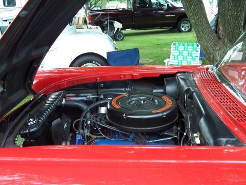 1975 Ford Ranchero - The engine bay of a red car is shown, with other vehicles and outdoor seating visible in the background.
