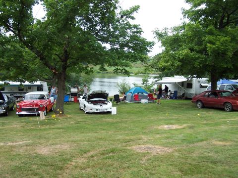 1975 Ford Ranchero - A gathering of vehicles and camping equipment is set up by a tranquil lake on a cloudy day.
