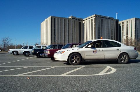 1991 Ford LTD Crown Victoria - A row of vehicles sits parked in a lot with large office buildings in the background under a clear blue sky.
