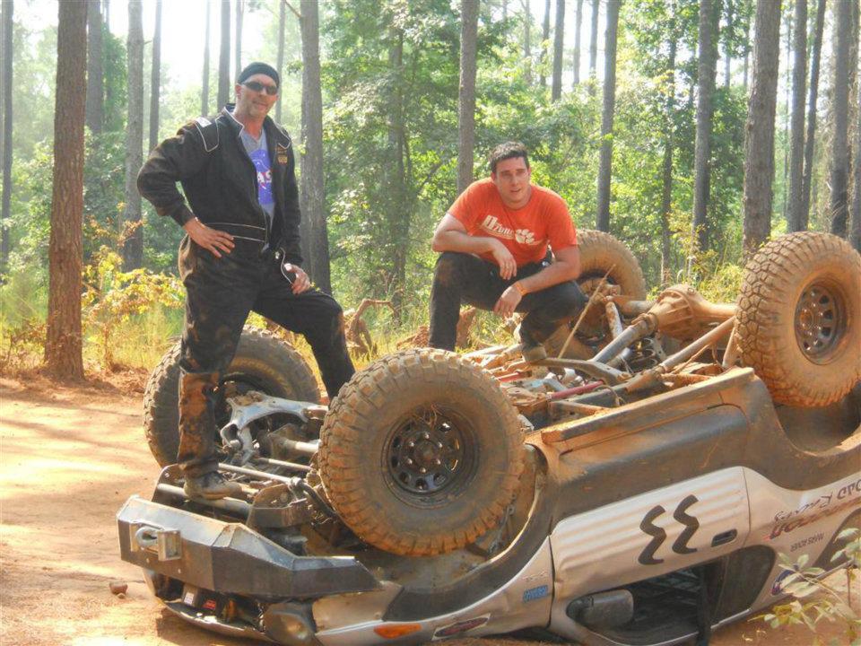 Overturned silver off-road vehicle with two men standing and crouching nearby
