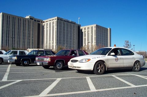 1991 Ford LTD Crown Victoria - Several police sedans are parked in a lot in front of a large office building.