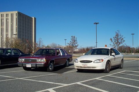 1991 Ford LTD Crown Victoria - Two sedans, a maroon 1991 Ford Crown Victoria LTD and a white sedan, are parked in a lot.
