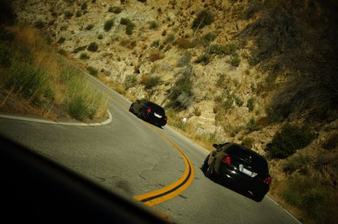 2003 Ford Crown Victoria - Two dark vehicles round a curve on a canyon road with rocky brush-covered slopes.
