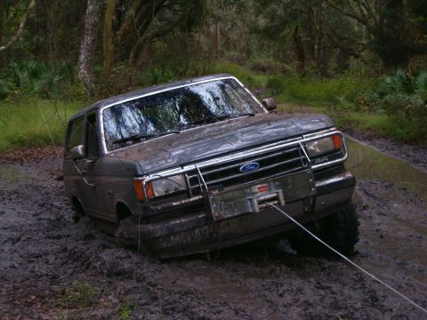 1991 Ford Bronco - A mud-caked full-size Ford Bronco buried to the axles in a deep mud hole on a wooded trail, leaning hard to the driver side with the front…