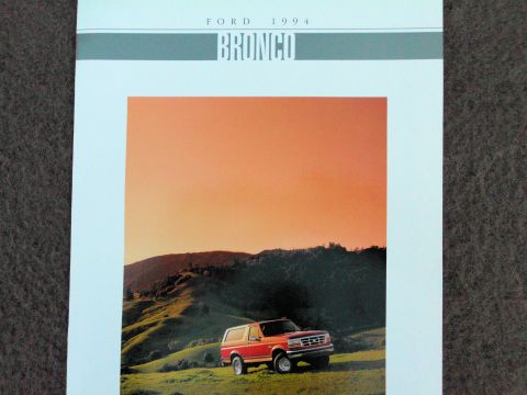 1994 Ford Bronco - Cover of a Ford 1994 Bronco sales brochure photographed on gray carpet, with 'FORD 1994' and a large 'BRONCO' wordmark over a teal band. Th…