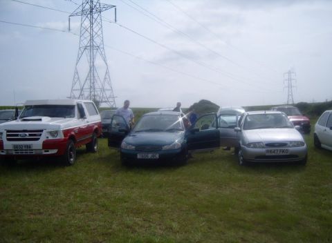 1989 Ford Bronco - A group of vehicles are gathered in a field under a cloudy sky with power lines overhead.
