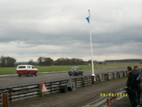 1989 Ford Bronco - Two vehicles, a red and white SUV and a dark sedan, are on a race track under a cloudy sky.