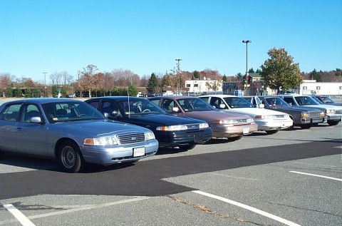 1991 Ford LTD Crown Victoria - A row of Ford Crown Victoria sedans, including a blue model in the foreground, are parked in a lot under a clear blue sky.