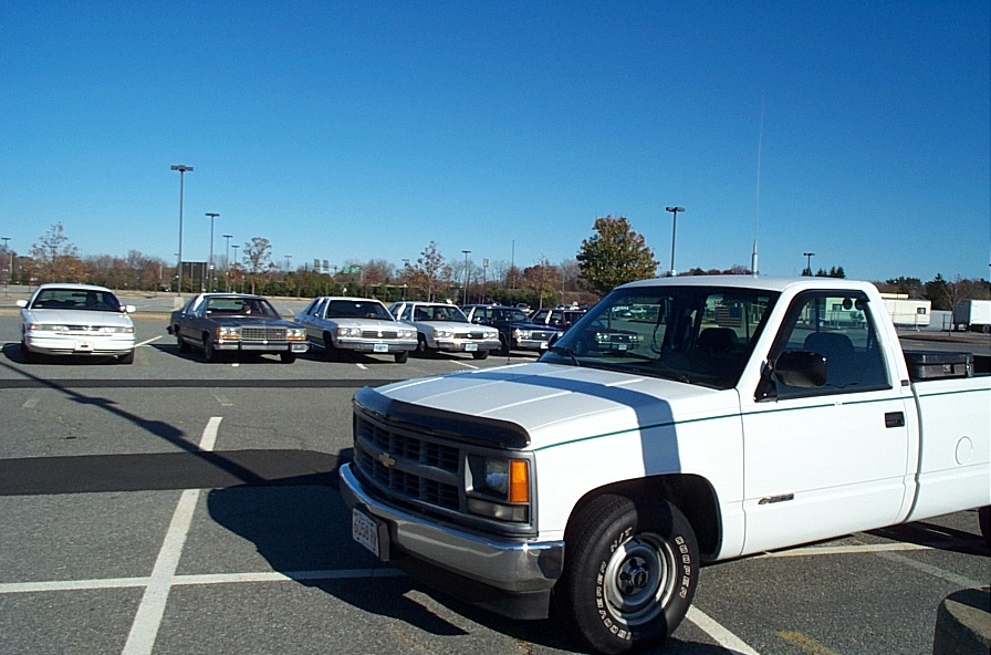 White pickup truck in foreground, several sedans parked in background under a clear blue sky