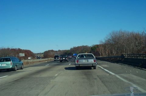 1991 Ford LTD Crown Victoria - A silver sedan travels down a highway on a clear day, with trees lining the road.