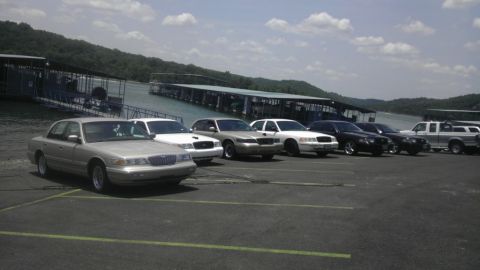 2000 Mercury Grand Marquis - A line of sedans and trucks are parked in a lot by a lake with a marina in the background.